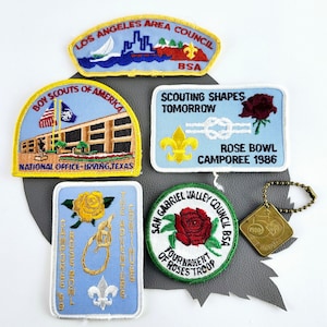 May include: Five embroidered patches for Boy Scouts of America. The patches are blue, white, and yellow with red roses, a sailboat, and a building. The text on the patches includes "Los Angeles Area Council", "Boy Scouts of America", "National Office-Irving, Texas", "Scouting Shapes Tomorrow", "Rose Bowl Camporee 1986", "San Gabriel Valley Council BSA", and "Tournament of Roses Troop".