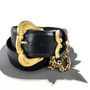 Puede incluir: Cintur&oacute;n de cuero negro con una hebilla grande dorada y detalles decorativos. El cintur&oacute;n presenta un detalle de cadena dorada colgando del lateral. La hebilla tiene un dise&ntilde;o ornamentado y vintage.