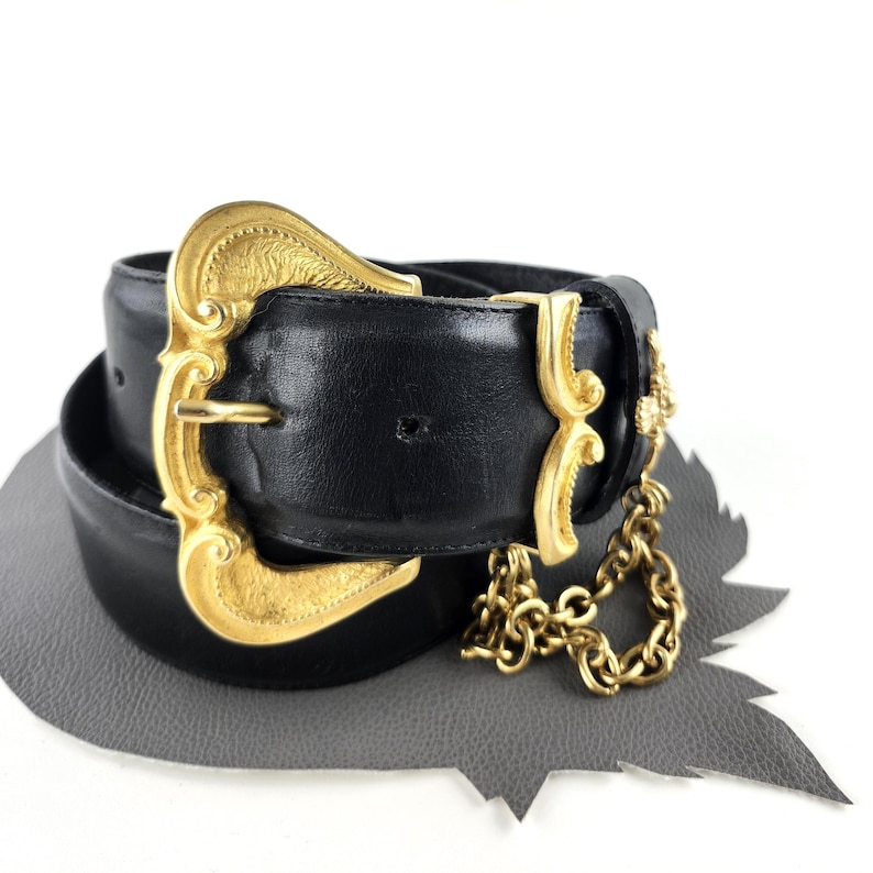 Puede incluir: Cintur&oacute;n de cuero negro con una hebilla grande dorada y detalles decorativos. El cintur&oacute;n presenta un detalle de cadena dorada colgando del lateral. La hebilla tiene un dise&ntilde;o ornamentado y vintage.