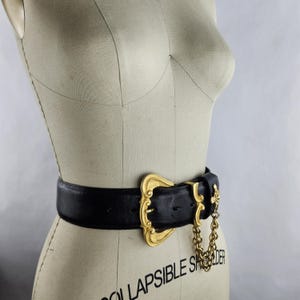 Puede incluir: Un cintur&oacute;n de cuero negro con una hebilla dorada grande y ornamentada. La hebilla tiene un dise&ntilde;o decorativo y un detalle de cadena. El cintur&oacute;n se muestra en un maniqu&iacute;.