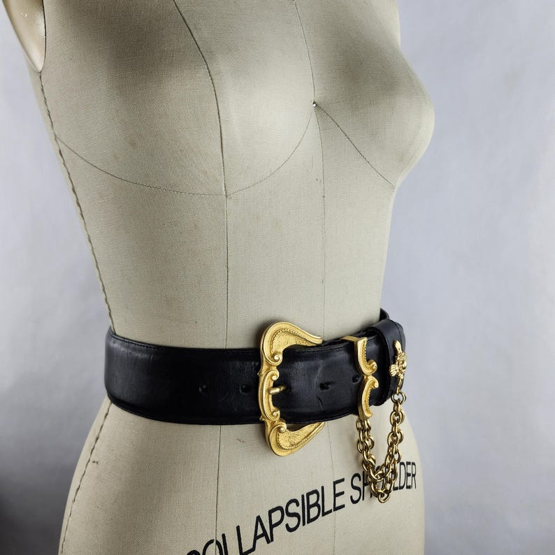 Puede incluir: Un cintur&oacute;n de cuero negro con una hebilla dorada grande y ornamentada. La hebilla tiene un dise&ntilde;o decorativo y un detalle de cadena. El cintur&oacute;n se muestra en un maniqu&iacute;.