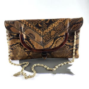 以下が含まれることがあります： 茶色と黒のヘビ革模様が特徴の長方形のクラッチバッグ。バッグのフロントフラップには半円形のダークブラウンの樹脂製ハンドルが組み込まれており、側面にはゴールドトーンの金属製チェーンショルダーストラップが付いています。