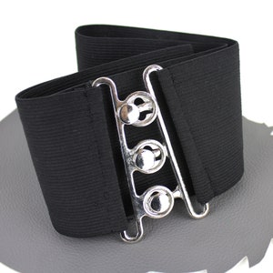 Könnte beinhalten: Ein schwarzer Gummiband mit einer silbernen Schnalle mit drei verstellbaren Schlaufen. Der Gürtel ist breit und hat eine glatte, gewebte Textur.