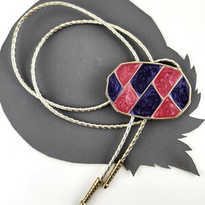May include: A vintage bolo tie with a gold-tone braided cord and decorative slide. The slide is an oval shape with a red, blue, and gold geometric design. The tie has gold-tone tips.