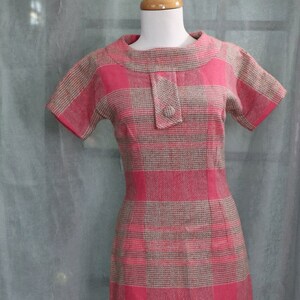 May include: A pink and brown plaid dress with a short sleeve and a round neckline. The dress has a button closure at the neck.