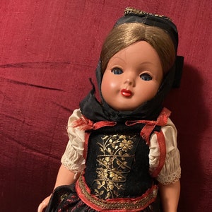May include: A vintage doll dressed in traditional attire. The doll has blue eyes, red lips, and brown hair. She wears a black bodice with gold embroidery, a black skirt with red and gold floral patterns, and a black head covering. The doll's sleeves are cream-colored with lace trim.