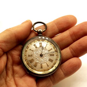 May include: An antique pocket watch with a silver-tone case and an ornate face. The watch has Roman numerals and gold-tone detailing. The watch is held in a hand, showing its size and design. The watch face has a diameter of approximately 4 cm.