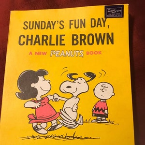 May include: A yellow book cover with the title "SUNDAY'S FUN DAY, CHARLIE BROWN" and the text "A NEW PEANUTS BOOK". The cover features a cartoon illustration of Charlie Brown, Snoopy, and Lucy dancing.