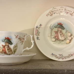 May include: A white teacup, saucer, and plate set featuring illustrations of Benjamin Bunny from Beatrix Potter. The teacup and plate have floral and bunny designs. The plate has the text "Benjamin Bunny" and "Beatrix Potter".
