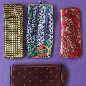 May include: Four different eyeglass cases. One is gold with a textured surface, one is blue and white with a floral pattern, one is red with a gold pattern and the text "Edgar Simons", and one is brown with a gold pattern.