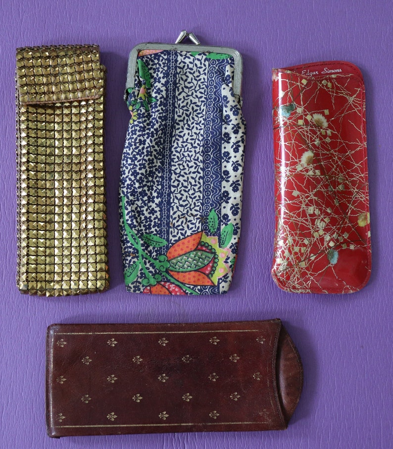 May include: Four different eyeglass cases. One is gold with a textured surface, one is blue and white with a floral pattern, one is red with a gold pattern and the text "Edgar Simons", and one is brown with a gold pattern.