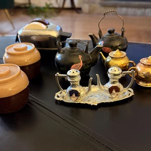 May include: A collection of vintage kitchenware, including two brown and tan salt shakers, several small teapots in gold and black, and a silver salt and pepper set with a decorative tray. A chrome toaster is also visible.
