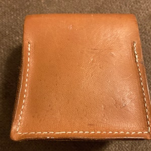 May include: A small, square, tan leather wallet with a flap closure. The edges are stitched with white thread. The wallet appears to be new and unused.