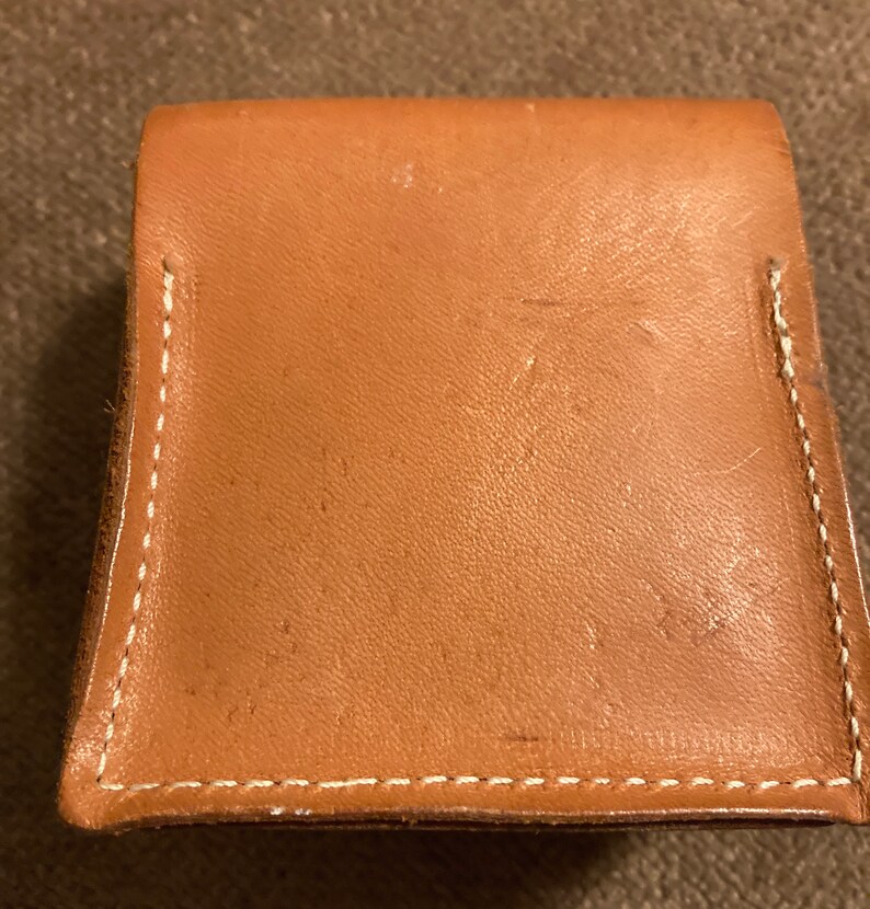 May include: A small, square, tan leather wallet with a flap closure. The edges are stitched with white thread. The wallet appears to be new and unused.