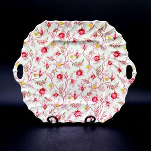 May include: A white ceramic serving tray with a scalloped edge and two handles. The tray is decorated with a floral pattern of red roses, yellow buds, and green leaves on a network of pink vines.