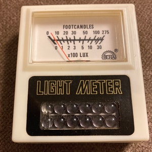 May include: A vintage light meter with a white plastic casing. The meter has a black and gold label that reads "LIGHT METER." The meter's gauge displays "FOOTCANDLES" and "x100 LUX."