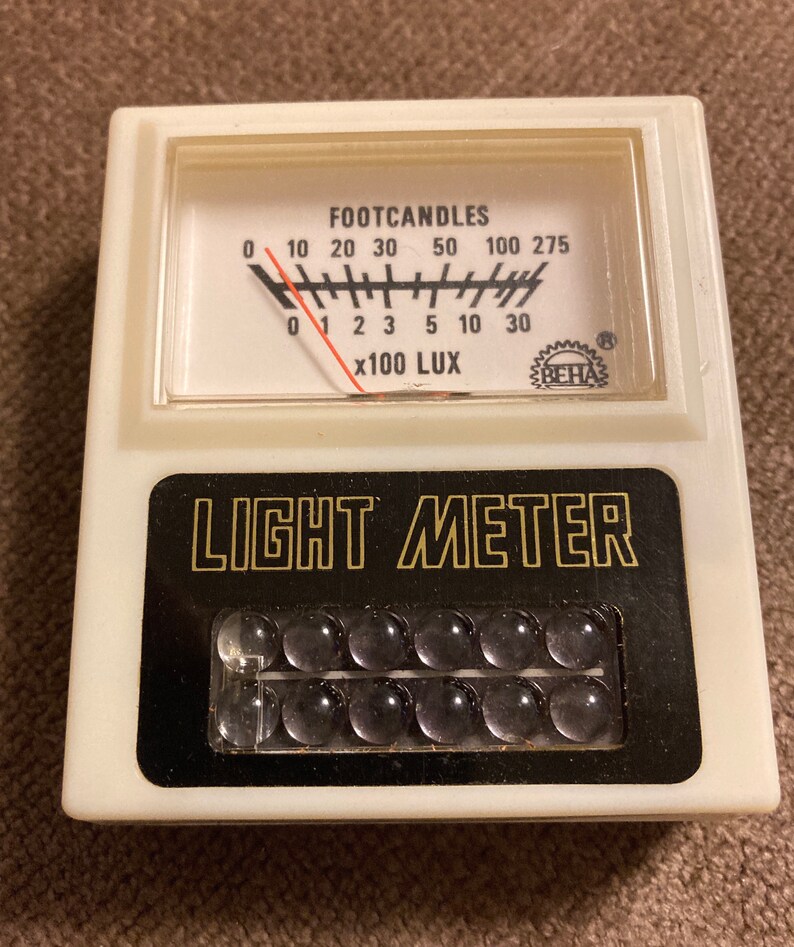 May include: A vintage light meter with a white plastic casing. The meter has a black and gold label that reads "LIGHT METER." The meter's gauge displays "FOOTCANDLES" and "x100 LUX."