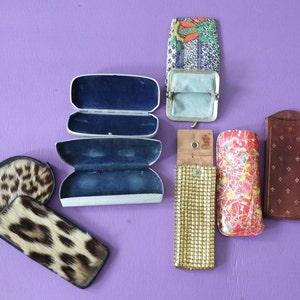 May include: Assortment of vintage eyeglass cases in various colors and patterns.  The cases are made of different materials, including leather, fabric, and metal.  Some of the cases have a floral design, while others have a geometric pattern.