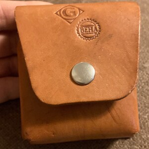 May include: A small, square, light brown leather pouch with a flap closure secured by a silver snap. The pouch has embossed logos, including a diamond-shaped design with a "G" and a circular emblem with "BHHA".
