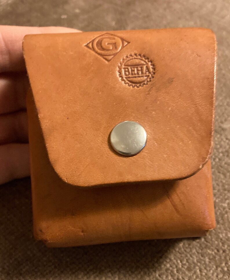 May include: A small, square, light brown leather pouch with a flap closure secured by a silver snap. The pouch has embossed logos, including a diamond-shaped design with a "G" and a circular emblem with "BHHA".