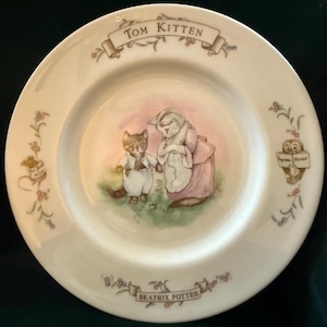 May include: A white ceramic plate featuring a watercolor illustration of two anthropomorphic cats. The plate is decorated with floral accents and text that reads "Tom Kitten" and "Beatrix Potter."