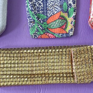 May include: Four different eyeglass cases. One is silver with a textured pattern, one is red with a gold pattern, one is blue and white with a floral pattern, and one is gold with a grid pattern.