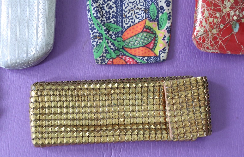 May include: Four different eyeglass cases. One is silver with a textured pattern, one is red with a gold pattern, one is blue and white with a floral pattern, and one is gold with a grid pattern.