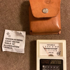 May include: A vintage light meter with a white face, black text, and a needle indicator. The meter reads in footcandles and lux. Includes a brown leather case and a paper with text: "GREENLEE/BEHA LIGHT METER TABLE OF ILLUMINATION LEVELS."