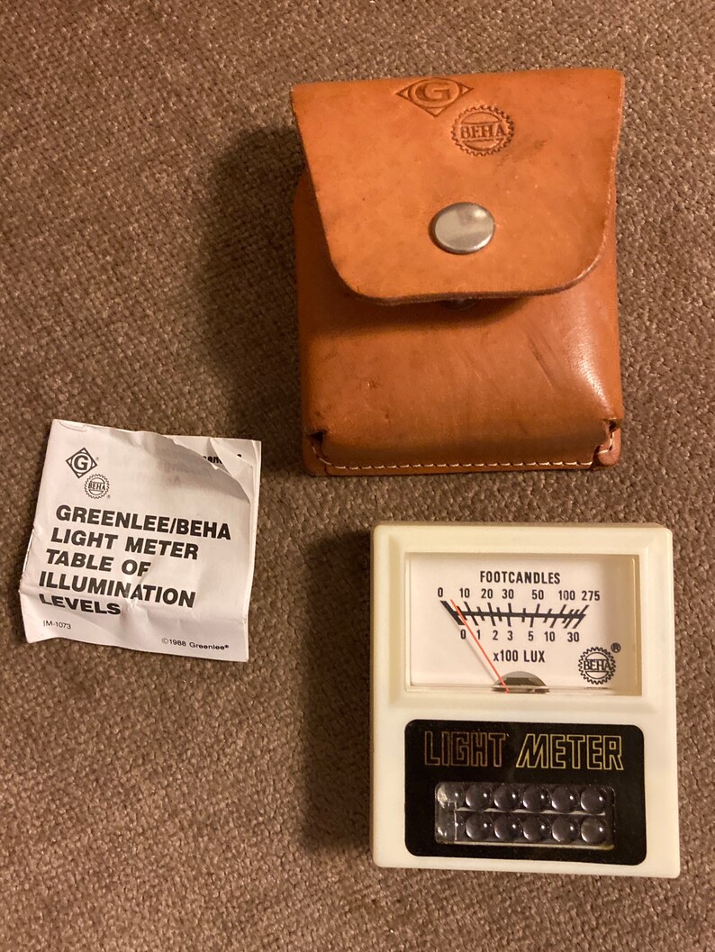 May include: A vintage light meter with a white face, black text, and a needle indicator. The meter reads in footcandles and lux. Includes a brown leather case and a paper with text: "GREENLEE/BEHA LIGHT METER TABLE OF ILLUMINATION LEVELS."