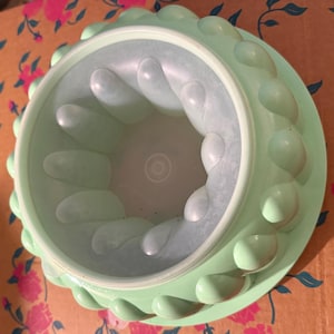 May include: A vintage, light green gelatin mold with a circular design. The mold features a series of teardrop-shaped bumps around the outer edge and interior. The center has a circular indentation. This kitchenware item is made of plastic.