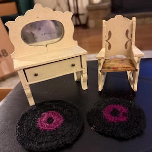 May include: A collection of miniature dollhouse furniture, including a cream-colored vanity with a mirror, a rocking chair, and two black crocheted coasters with a purple center. The items are arranged on a dark surface.