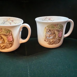 May include: Two white ceramic mugs with a whimsical illustration of rabbits in a forest setting. The mugs have a curved handle and a floral design near the rim. The mugs are on a dark green surface.