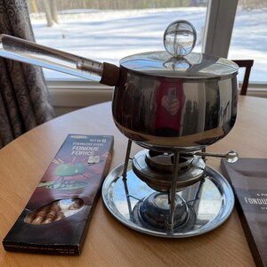 May include: A stainless steel fondue pot with a clear glass handle and lid knob. The set includes a box of six stainless steel fondue forks. The pot sits on a metal stand with a burner.