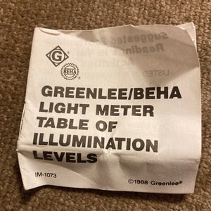May include: A crumpled white paper with the text "GREENLEE/BEHA LIGHT METER TABLE OF ILLUMINATION LEVELS." The paper has the "G" and "BEHA" logos. The text "IM-1073" and "&copy;1988 Greenlee&reg;" are also visible.