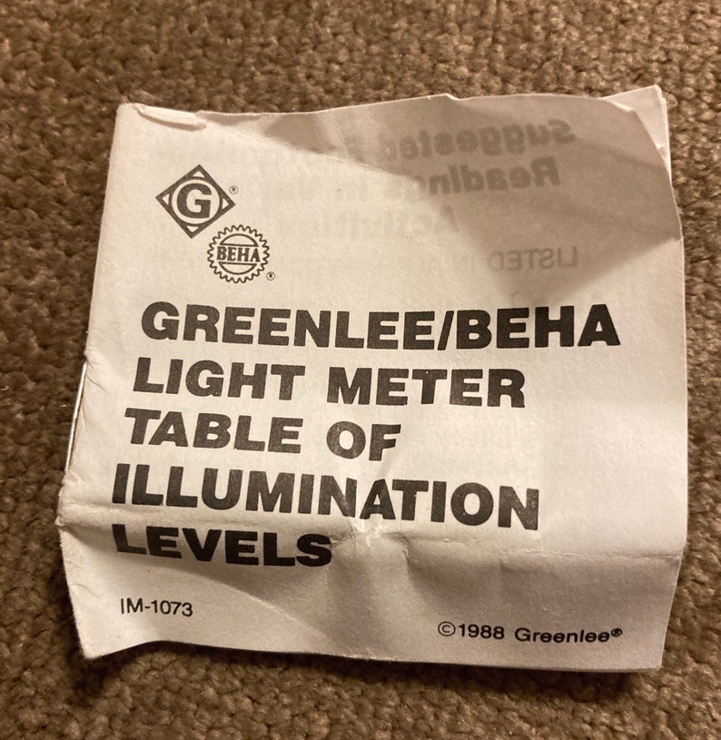 May include: A crumpled white paper with the text "GREENLEE/BEHA LIGHT METER TABLE OF ILLUMINATION LEVELS." The paper has the "G" and "BEHA" logos. The text "IM-1073" and "&copy;1988 Greenlee&reg;" are also visible.