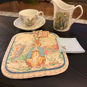 May include: A cream-colored Peter Rabbit themed tea cup and saucer set, a matching creamer, a pot holder, and a notepad. The pot holder features Peter Rabbit in a blue jacket. The notepad has the words "Peter Rabbit" printed on it.