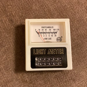 May include: A vintage light meter with a white body and black accents. The meter displays "FOOTCANDLES" and "x100 LUX" with a needle indicating light intensity. The text "LIGHT METER" is prominently displayed.