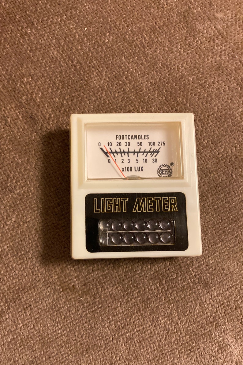 May include: A vintage light meter with a white body and black accents. The meter displays "FOOTCANDLES" and "x100 LUX" with a needle indicating light intensity. The text "LIGHT METER" is prominently displayed.