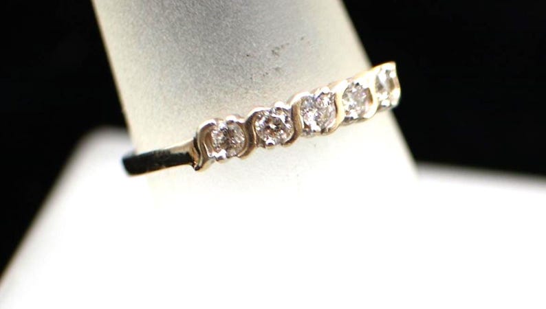 以下が含まれることがあります： 一列に並んだ5つの丸い透明な石が特徴のゴールドリング。金属製のバンドは滑らかで光沢のある仕上げで、石は個別の金属製の台座にセットされており、リングの上部にスカラップ状の縁取りが施されています。