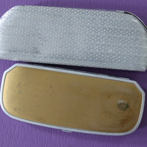 May include: Two vintage eyeglass cases. The top case is silver with a diamond pattern. The bottom case is gold with a worn surface.