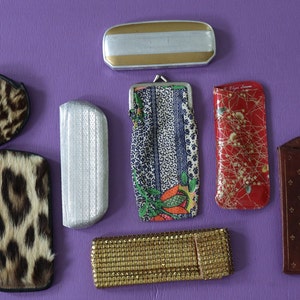May include: Assortment of vintage eyeglass cases in various colors and patterns, including leopard print, silver, gold, red with floral design, and brown with a fleur-de-lis pattern.