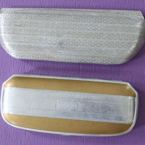 May include: Two vintage metal eyeglass cases. The top case is silver with a textured pattern. The bottom case is gold with a silver band.