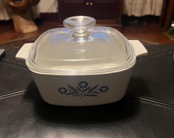 May include: White ceramic casserole dish with a glass lid and a blue floral design. The dish has a square shape and is perfect for baking and serving.