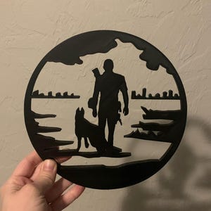 May include: Black silhouette of a person and a dog standing on a beach with a cityscape in the background. The silhouette is cut out of a round piece of wood.