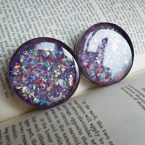 May include: Two round, clear, purple resin plugs with iridescent glitter embedded inside. The plugs are on a book with text.