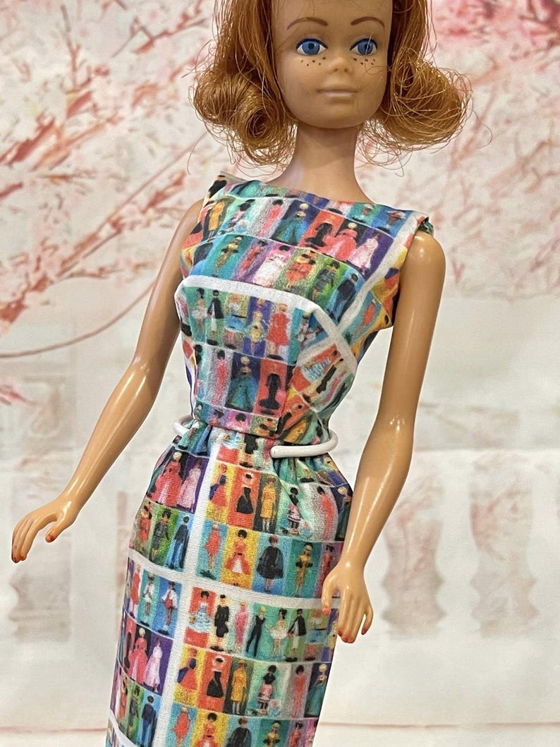May include: A vintage doll wearing a colorful, patterned dress. The sleeveless dress features a white belt and a print of various figures in different outfits. The doll has red hair and blue eyes.