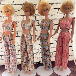 May include: Four vintage Barbie dolls wearing matching two-piece outfits. The outfits feature various patterns, including a collage of images, zebra stripes, and a pink print with the Barbie logo. Each doll has a different hairstyle and is standing on a white base.
