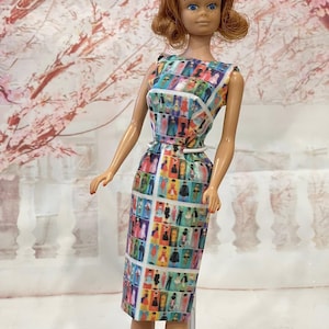 May include: A vintage doll with red hair and blue eyes, wearing a colorful dress with a grid pattern of figures. The dress has a white belt and a sleeveless design. The doll is standing in front of a soft, blurred background.