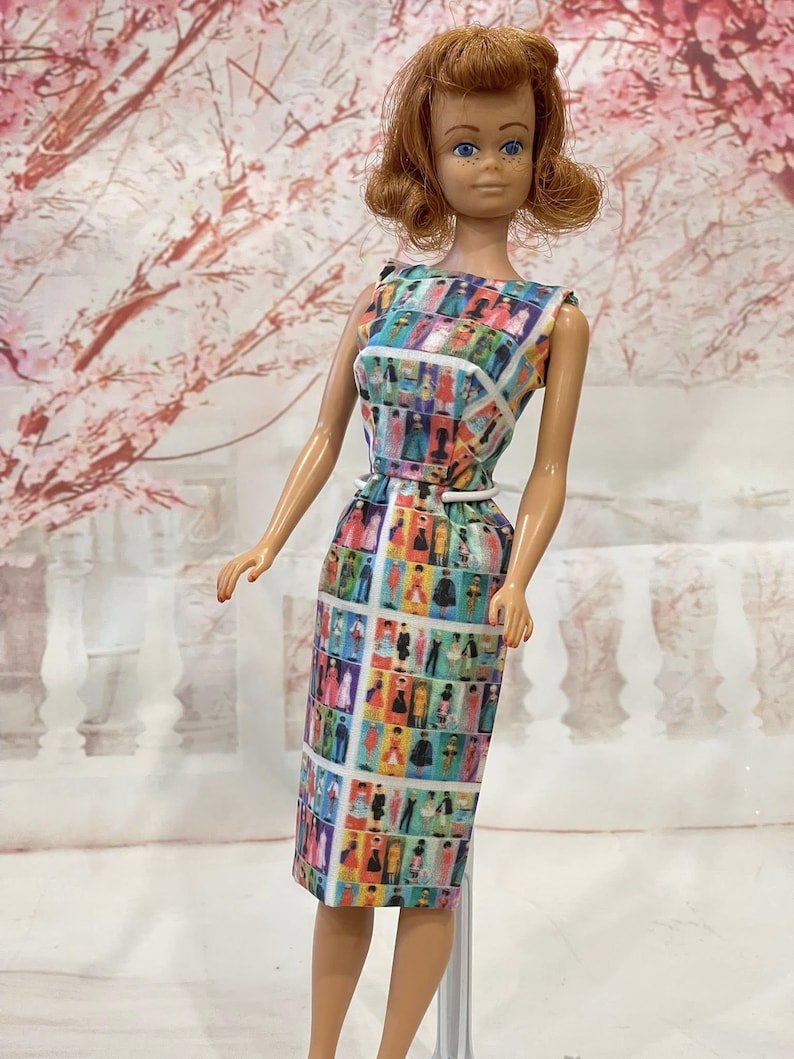 May include: A vintage doll with red hair and blue eyes, wearing a colorful dress with a grid pattern of figures. The dress has a white belt and a sleeveless design. The doll is standing in front of a soft, blurred background.