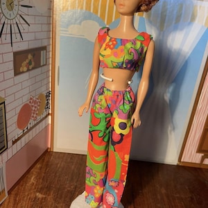 May include: A vintage doll in a vibrant two-piece outfit with a floral design in red, green, yellow, and pink. The doll has red hair and stands on a white base. The background features a retro interior design.